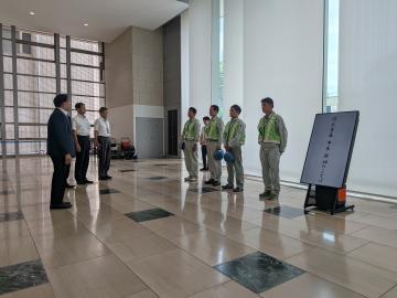 広いロビーで制服の作業員数人がヘルメットを持ち整列して立ち対面にスーツ姿の数人が並び右側に縦型の電子看板が置かれている