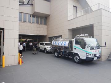 建物の入口前に給水タンクを載せたトラックや白い車が停まり数名の作業員が出入りしている