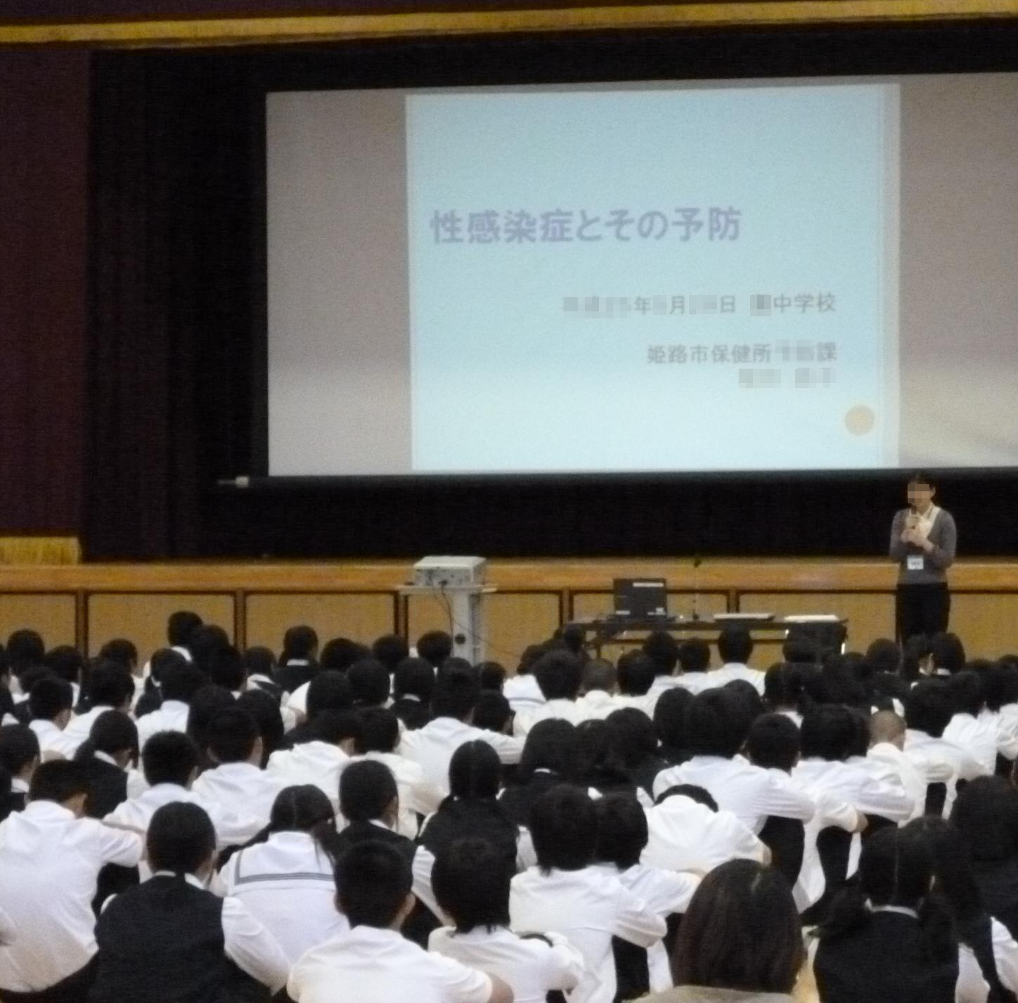 体育館のような大きなホールで多数の生徒が座ってスクリーンを見て壇上の講師がプレゼンテーションをしている様子