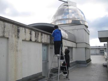 生徒1人が脚立にのぼり、もう1人は脚立を押さえて天文台の塗装をするようす