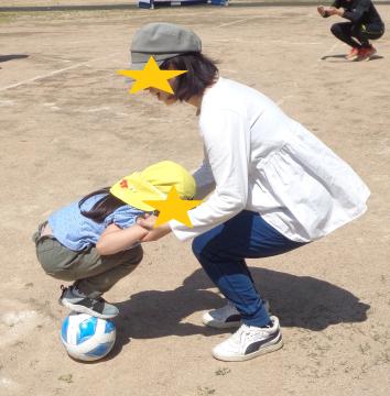 お家の人に支えてもらいサッカーボールの上でしゃがむ