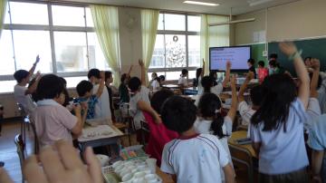 教室内で児童がクイズ大会をしています。3択クイズにたくさんの手が挙がっています。