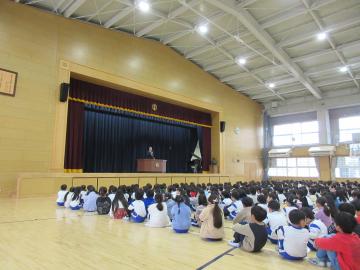 体育館に児童たちが整列して座り、壇上に先生が立って話をしている終業式の様子