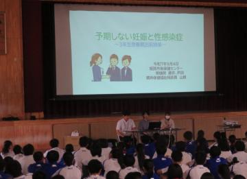 体育館でスクリーンを見ながら講師の話を聞いている生徒たちの様子