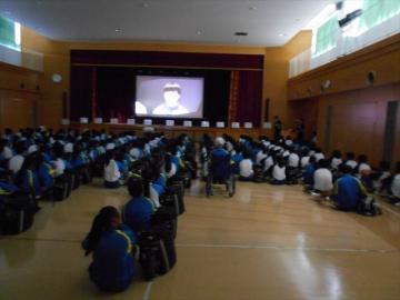 体育館で壇上に設置されたモニターに映された映像で学習をしている生徒たちの様子