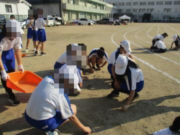 大勢の生徒が陸上トラックのレーンの周りの草を抜いています。