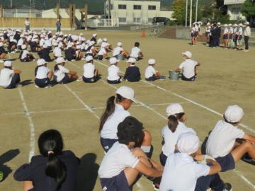 校庭に座りたてわり班ごとに説明を聞く児童たちの様子