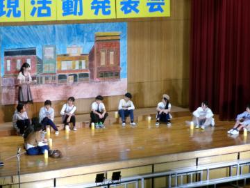 体育館の壇上で、5年生と6年生の児童たちがお芝居の中で語っている様子