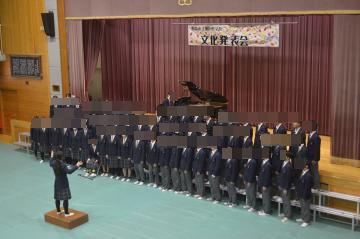 体育館の舞台の上で伴奏者がピアノを演奏し、舞台の前で三年生全員が3列に並び旅立ちの日にを歌っています。指揮者は少し離れた位置に立ち、指揮をしている様子