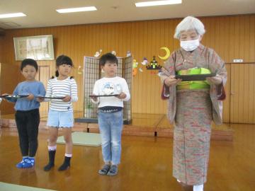 遊戯室で盆に和菓子をのせて、着物を着た講師を見ながら運ぶ園児たちの様子