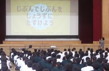 体育館で壇上のプロジェクターに映された画面を観ながら講師の話を聴く生徒たちの様子