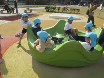 公園内の回転する遊具に子供たちが座って乗り、一人が外側から回している園児たちの様子