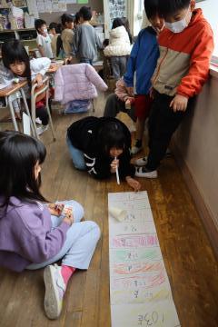 教室の床に転がった紙コップを児童がストローで吹いています