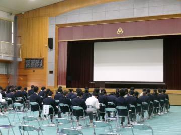 生徒が体育館の椅子に座って、体育館舞台前にいる学年主任の話を聞いています。