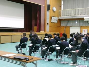 体育館の中央で学年主任が進路について説明しています。生徒は椅子に座って説明を聞いています。