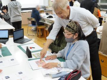 椅子に座って記事を見ている女性に、三好先生が後ろからアドバイスをしている様子