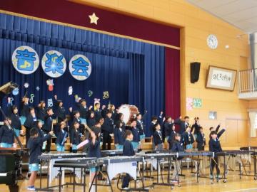 3年生が体育館の壇上とその前に整列して楽器の合奏をしながら左拳を上に突き上げている
