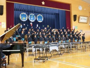 3年生が体育館の壇上に整列して合唱をしながら左拳を上に突き上げている