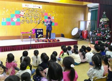 地域のおじいさんが体育館の広間の壇上にあがり、披露してくれるマジックショーを、床に座って見る園児たちの様子