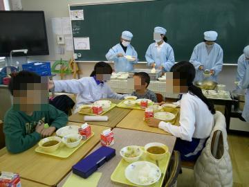 教室で給食の配膳を背景に、4つの勉強机を向き合わせてグループにしで座っている様子