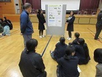 ポスターを提示したパネルの前に体育座りをして、高校生の説明を聞く児童・生徒の様子