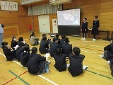 スクリーンの前に体育座りをして、高校生の説明を聞く児童・生徒の様子