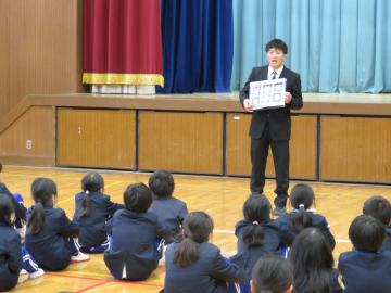 生活指導担当の先生が整列して座る児童たちの前に立ち、フリップをもって話をしている