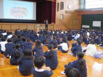 体育館に全校生徒が整列して座り、冬休みのくらしについての話を先生から聞くようす