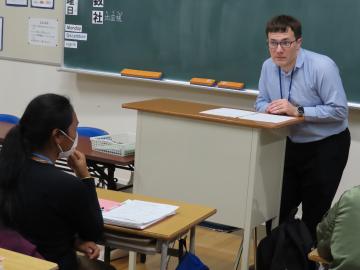 教室の教壇に立つ教師に向かって、生徒が冬休みの質問をしています。