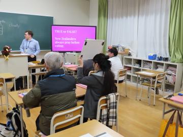 教室で教壇に立つ先生がニュージーランドのクイズを出して、生徒がホワイトカードを掲げてクイズに答えています。