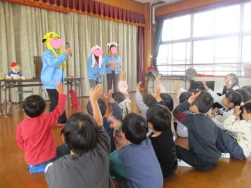 遊戯室内でマイクをもった講師に向かって挙手をしている子供たち