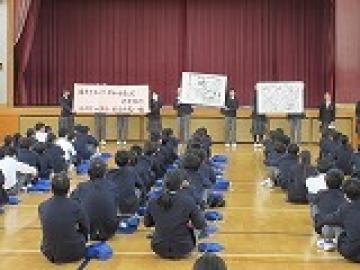 体育館の壇上前でスローガンを書いた紙と拡大した表紙絵2枚を掲げる実行委員の様子