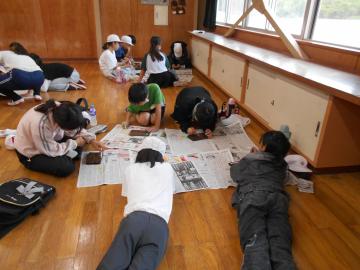 5年生が部屋の床に座ったり寝そべったりしながら、野外炊飯で作った焼き板に絵付けをしている様子