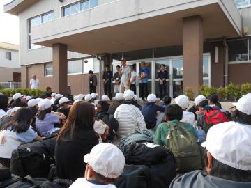 5年生が施設の玄関前に整列して座り、前に立つ先生の話を聞いている解散式の様子