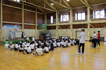 5年生が野外活動センターの体育館に整列して座り、前に立つスタッフの説明を聞く様子