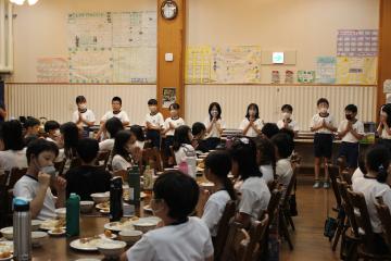 食堂で着席した生徒たちと、前に立つ数名の生徒が手を合わせて「いただきます」と言っている様子