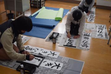 教室内で6年生が床に置かれた用紙に書初めをする様子