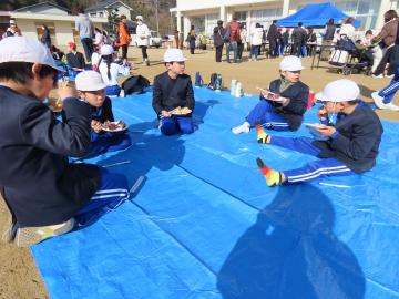 校庭でブルーシートに座りお餅を食べている児童たちの様子