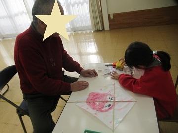 地域の方と教室のテーブルで凧つくりを教えてもらっているこども