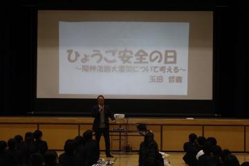 体育館で「ひょうご安全の日」と書かれたプロジェクターを映し、講師が立って震災学習の説明をするようす