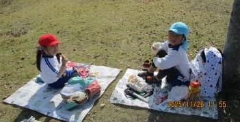 芝生の上にレジャーシートを敷いて、その上でピクニックをする園児たちの様子