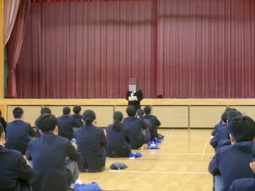体育館の舞台前中央で教員が受験当日のエピソードを語っています。生徒は体操座りをして話を聞いています。