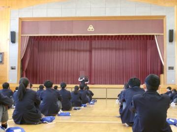教員が体育館舞台前中央で生徒に向けて受験についてと三学期の過ごし方について話しています。