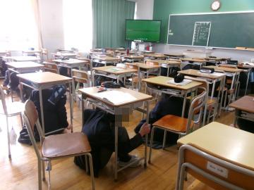 3年2組の生徒が、避難訓練のため一時的に机の下に避難しています。