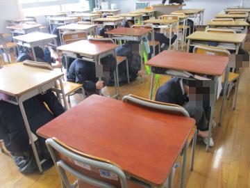 3年1組の生徒が、避難訓練のため一時的に机の下に避難しています。