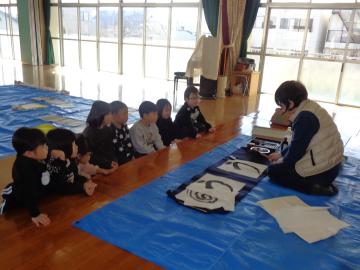 遊戯室で幼児が習字の先生から文字の書き方を教えてもらっている様子