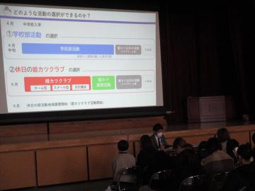 体育館のスクリーンを使い、参加者へ部活動について説明する先生の様子