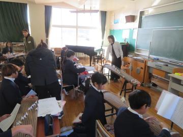 音楽室の教壇に立ち箏の音の出し方を5年生に説明する講師と、箏を触る生徒達