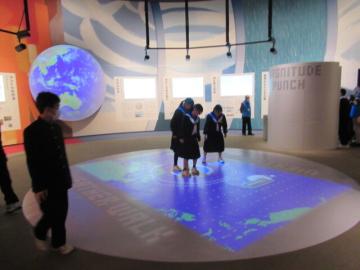 自然災害の体験コーナー　自然現象のメカニズムが床に映し出され、その上で床を見て自然現象のメカニズムを学ぶ生徒たち