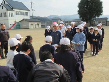 運動場で立ったまま、屋外スポーツクラブの紹介をする6年生の集団とその話を聞く3年生の集団